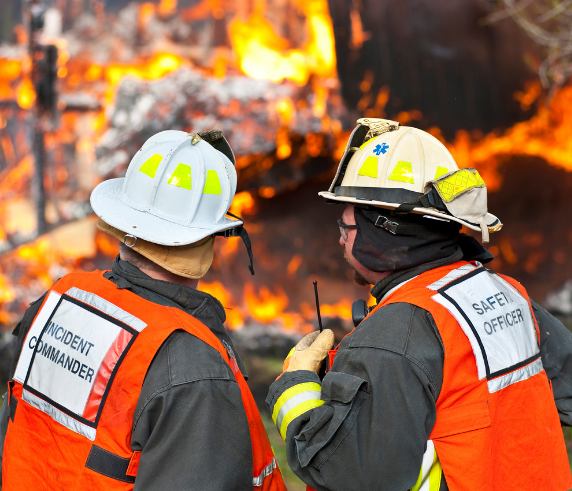 Two incident commanders communicating and assessing a fire with burning structure in the background.