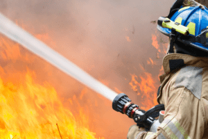 Fireman with a hose under his arm actively extinguishing a large wildfire in the background.