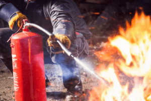 Man in fire safety clothing extinguishing a large fire with a red fire extinguisher in a burnt area.