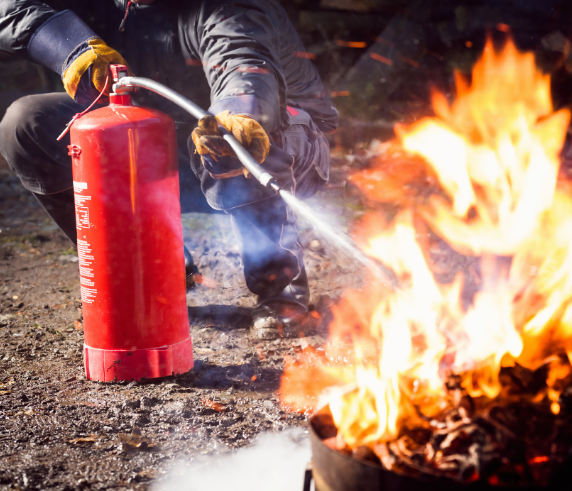 Man in fire safety clothing extinguishing a large fire with a red fire extinguisher in a burnt area.