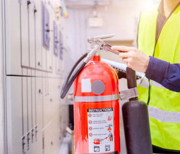 Person in PPE holding up a red fire extinguisher in a industrial workplace locker room.