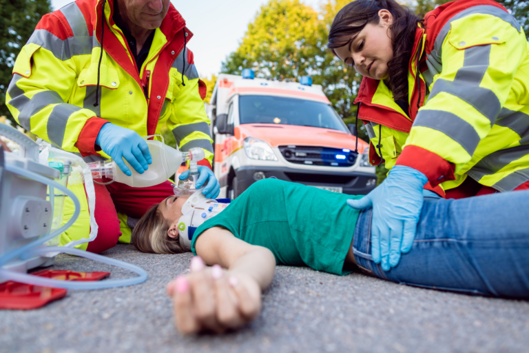 Two paramedics performing first aid to a woman laying on the floor wearing a neck brace with an ambulance vehicle in the background.