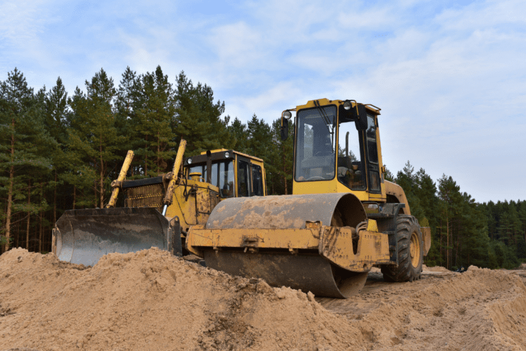 Conduct Roller Operations. A yellow roller and a bulldozer on a sandy construction site with a forest in the background