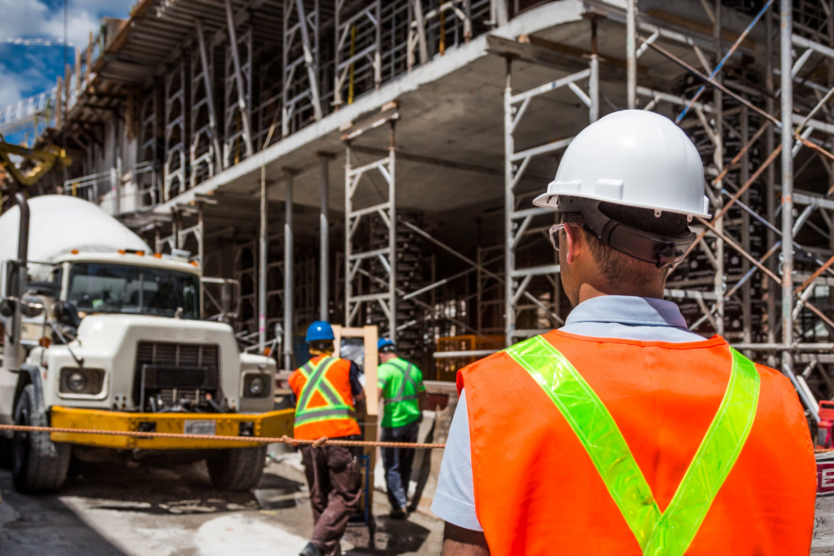 Worker standing to the left wearing HiVis and a white helmet looking at construction building scaffold and workers moving boxes on a trolley.