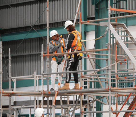 Two scaffolders wearing harnesses and white hard hats standing on a high-level scaffold looking down below.
