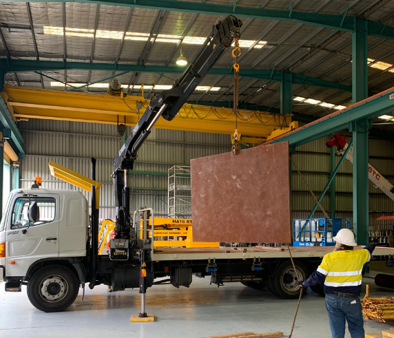 Operate a Vehicle Loading Crane. A vehicle loading crane lifting a large slab onto a truck in an industrial warehouse