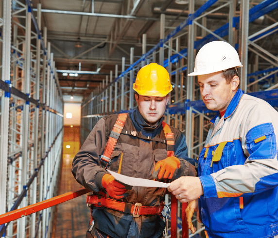 Work Safely and Follow WHS Policies and Procedures. Two workers in protective gear discussing safety protocols in a warehouse setting with scaffolding