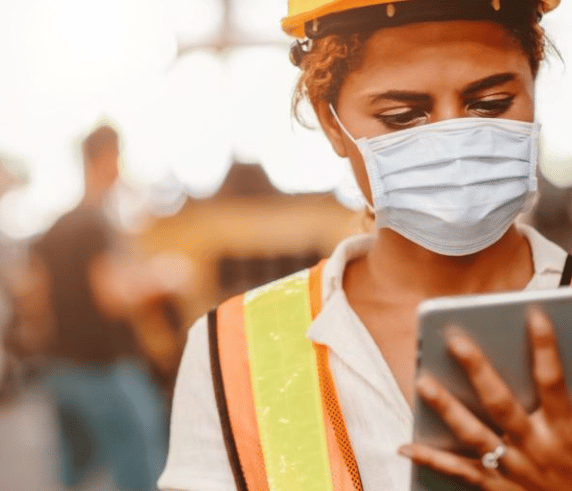 Female worker wearing a high visibility vest and white hardhat holding a electronic tablet.