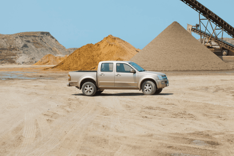 Parked beige Ute light vehicle in the middle of jobsite with mounds of sand and mining machinery in the background.