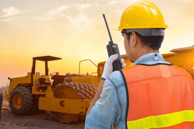 Work Safely and Follow WHS Policies and Procedures. Construction worker in high-visibility vest using a walkie-talkie on a worksite with heavy machinery during sunset