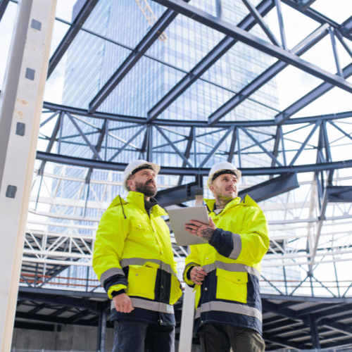 Two men wearing yellow Hi-Vis and white hardhats holding a electronic tablet standing under large construction.