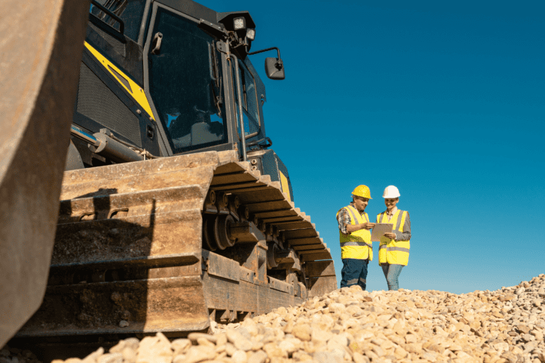 Statutory Supervisor WHS Risk Management. Two construction supervisors wearing safety gear and helmets reviewing documents beside a large tracked excavator on a gravel site