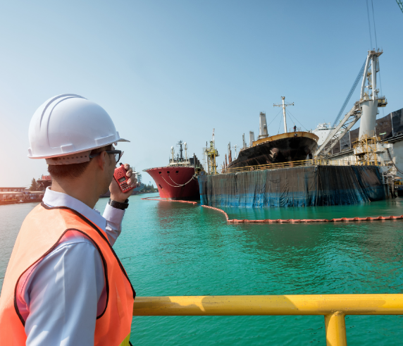 An individual in orange safety coveralls holding a clipboard and writing or reviewing notes in an industrial setting, possibly a factory or plant, with various pipes and valves in the background. The person’s face is obscured by a pixelated block for privacy, and there are bright yellow elements, likely safety barriers or parts of machinery, that stand out due to their color.