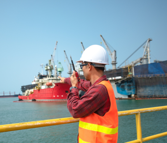 A person in safety gear standing on an offshore platform, looking towards a large red and black ship with cranes docked near the platform, under clear skies, indicating a maritime industrial setting or offshore construction and maintenance activities.