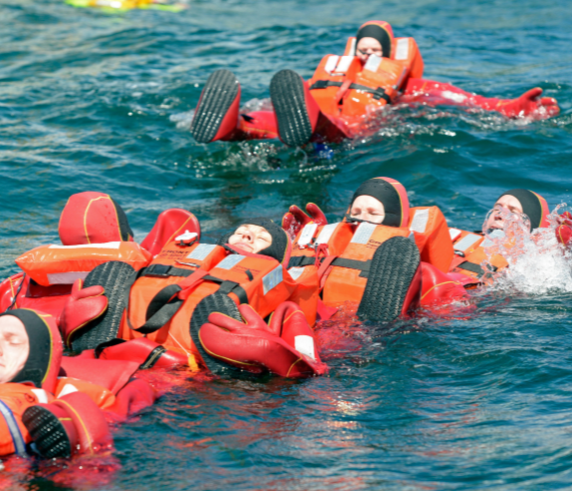 A group of individuals in the water wearing red life vests and white helmets, participating in a water survival training exercise. They are floating on their backs with their legs elevated, likely to keep afloat and conserve energy. In the background, there is an overturned bright orange raft with black tires attached to it, indicating a simulated emergency scenario for the training exercise.