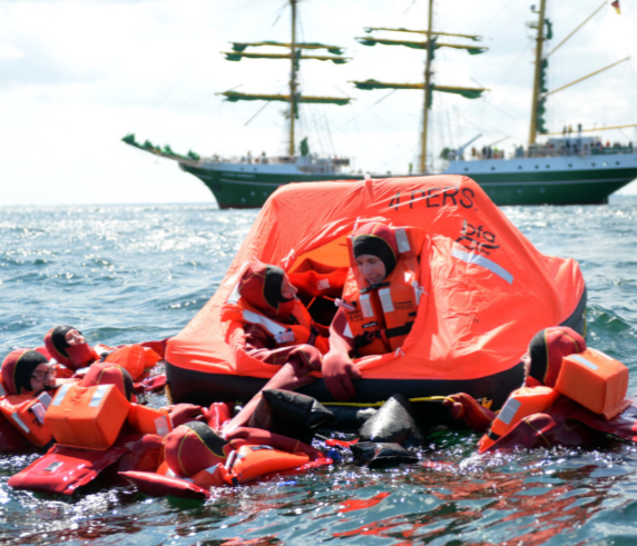 A group of individuals in life jackets huddled under an orange survival raft in the open sea, with a large ship with multiple masts and sails in the background, suggesting a maritime emergency drill or an actual distress situation.