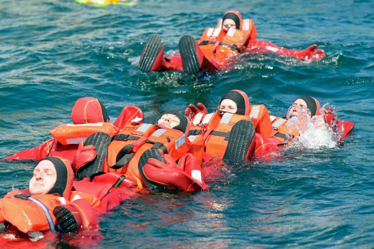 A group of people in bright orange survival suits and life vests are floating on their backs in the water. They are practicing survival techniques, possibly in a training exercise. The individuals are clustered together, maintaining a formation to stay afloat.