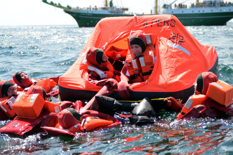 A group of people in bright orange survival suits and life vests are floating in the water, surrounding an orange inflatable life raft marked "4 PERS." Two individuals are inside the raft, with the others holding onto the edges. In the background, a green and white ship is visible on the horizon.