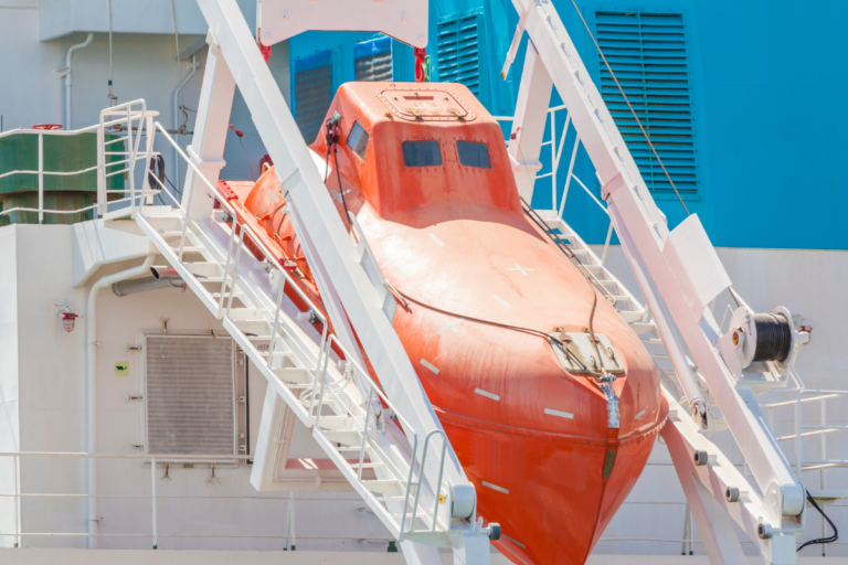 An orange freefall lifeboat is secured in its launching system on a ship. The lifeboat is positioned at an angle, ready for deployment. The background shows parts of the ship's structure, including stairs and blue ventilation panels.