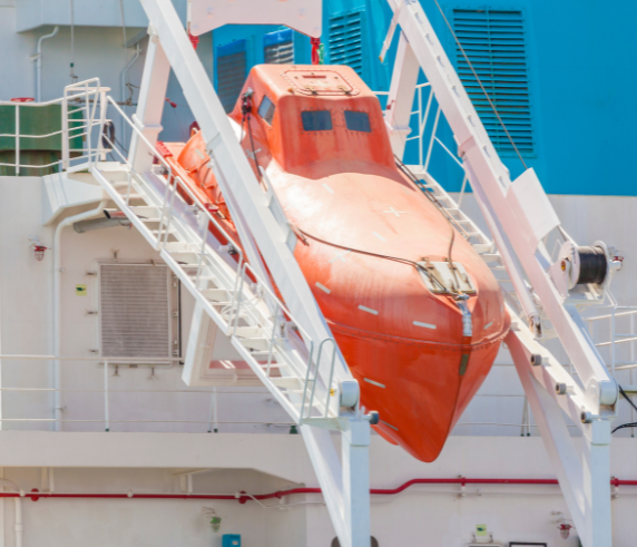 Freefall Lifeboat Survival Craft. An orange freefall lifeboat secured on the deck of a maritime vessel, ready for deployment. The lifeboat is positioned within a white launch frame against a backdrop of blue and white structural elements.