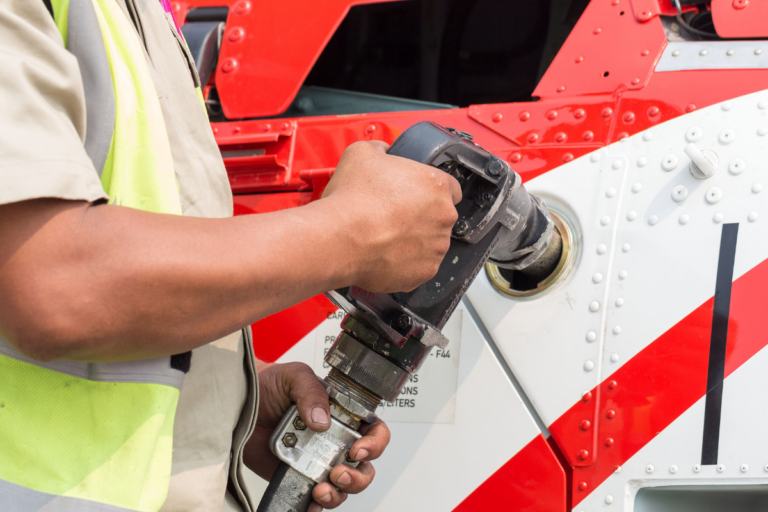 The image shows a close-up of a person refueling a helicopter. The individual is holding a fuel nozzle and inserting it into the helicopter's fuel tank. The helicopter's exterior is painted white with red stripes and visible rivets. The person refueling the helicopter is wearing a high-visibility vest, suggesting they are following safety protocols. The focus is on the refueling process, highlighting the careful and precise nature of this task.