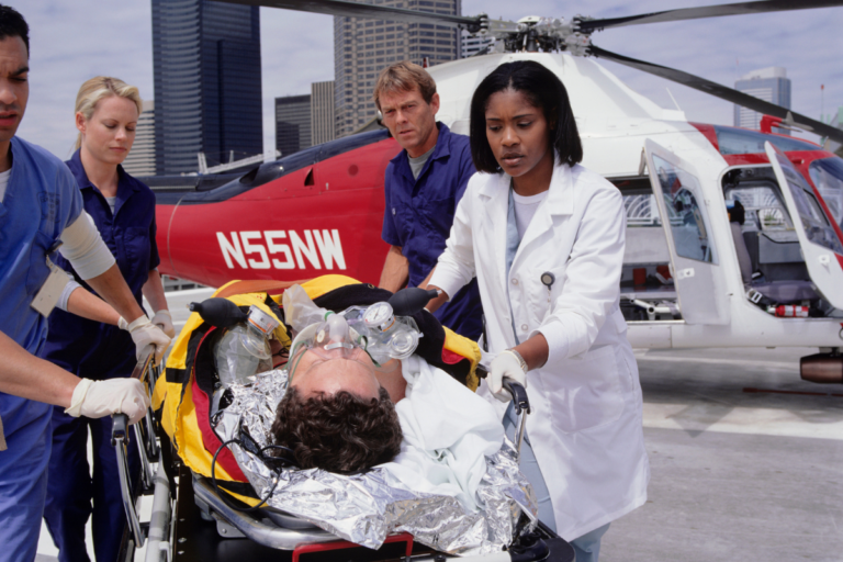 A medical team, including a doctor and nurses, is seen urgently transporting a patient on a stretcher from a red and white rescue helicopter marked "N55NW." The patient is receiving oxygen and other medical care, indicating a critical emergency. The background features tall city buildings, suggesting the scene is taking place on a rooftop helipad in an urban environment. The medical personnel are focused and professional, ensuring the patient receives immediate attention upon arrival.