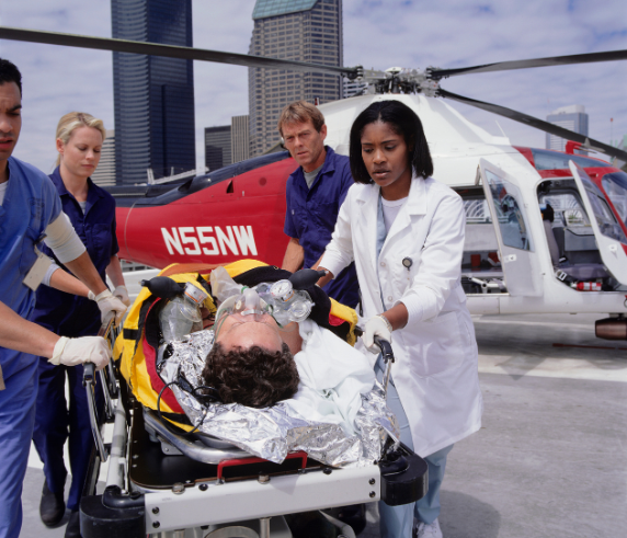 An emergency medical scene with a patient on a stretcher receiving attention from medical personnel, including individuals in blue scrubs and a white coat. A red helicopter with the registration number N55NW is visible in the background, indicating an air medical service. The setting appears to be an urban area with skyscrapers under a clear sky.