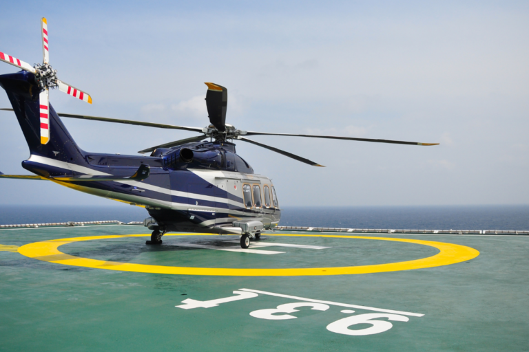 A blue and white helicopter is parked on a helideck marked with a large yellow circle and the number "9.3T." The helicopter's rotor blades are stationary, and the ocean is visible in the background, with clear blue skies overhead.