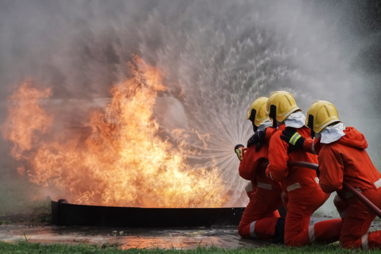 Four firefighters in full protective gear, including helmets and orange suits, are tackling a large fire. They are kneeling on the grass and using a hose to spray water at the intense flames, which creates a dramatic splash of water against the fire. The teamwork and coordination of the firefighters are evident in their focused effort to extinguish the blaze.