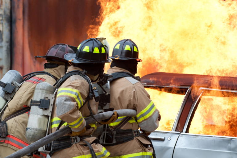 Image depicts three firefighters in full gear, including helmets and oxygen tanks, standing near a vehicle engulfed in flames. The intense orange and yellow fire is blazing behind them, indicating an active and dangerous firefighting scene. The focus on the firefighters suggests their role in emergency response and the importance of fire safety measures.