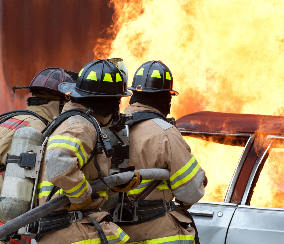 Image depicts three firefighters in full gear, including helmets and oxygen tanks, standing near a vehicle engulfed in flames. The intense orange and yellow fire is blazing behind them, indicating an active and dangerous firefighting scene. The focus on the firefighters suggests their role in emergency response and the importance of fire safety measures.