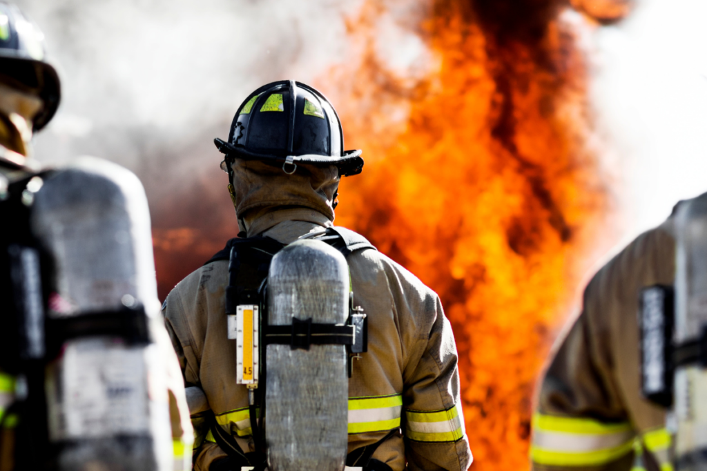 Three firefighters are seen from behind, standing in front of a large blaze. They are wearing full firefighting gear, including helmets, protective suits, and breathing apparatuses on their backs. The fire is intense, with bright orange flames and thick smoke billowing upward. The scene captures the urgency and danger of firefighting.