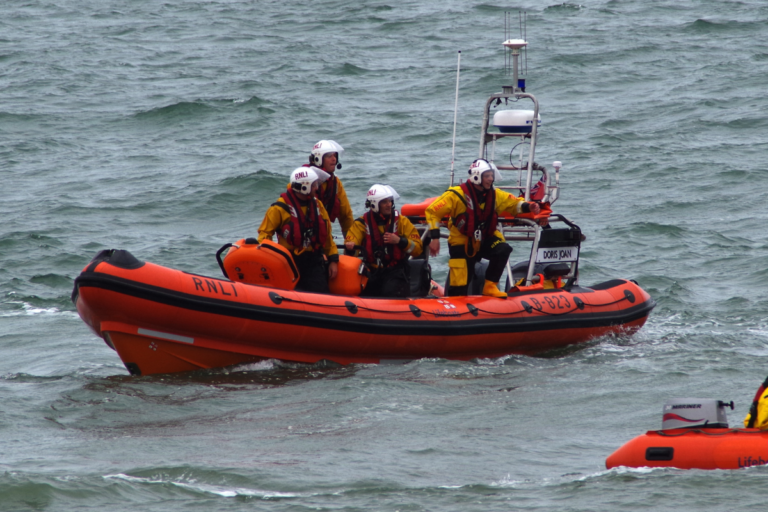 A team of rescue personnel in yellow and black protective gear, labeled "RNLI," are on an orange lifeboat named "DORIS JOAN." They are navigating through choppy waters, equipped with helmets and life jackets, appearing ready for a rescue operation. Another smaller orange rescue boat is partially visible in the lower right corner.