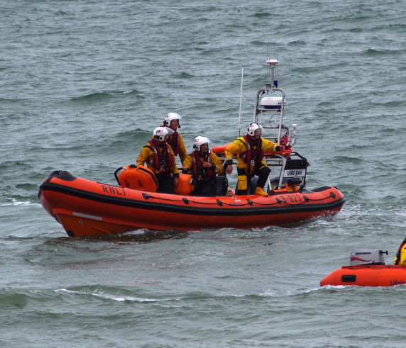 A group of individuals wearing bright yellow life jackets and helmets are aboard an orange rigid inflatable boat (RIB) on choppy sea waters. The boat is equipped with various navigational and safety equipment, indicating a focus on personal survival techniques at sea.