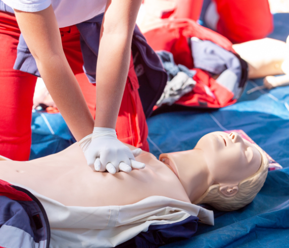 A person performing CPR on a training mannequin. The individual is shown pressing down on the mannequin’s chest with interlocked hands to demonstrate the chest compression aspect of cardiopulmonary resuscitation. The setting appears to be an outdoor training session, as suggested by natural lighting and a blurred background indicating an open environment. This image is relevant for educational purposes, illustrating the technique of administering CPR, which is a critical emergency procedure that can save lives.