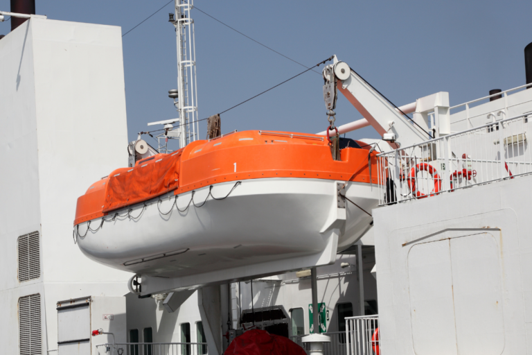 A bright orange and white enclosed lifeboat is secured on the deck of a ship, held in place by a swing-arm davit system. The lifeboat is ready for deployment, and various safety equipment, such as life rings, are visible on the ship's railing. The ship's structure, including ladders and ventilation, can be seen in the background against a clear sky.