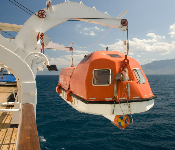 An orange lifeboat is suspended above a white deck by a swing arm davit, which is a type of crane mechanism used for lowering and raising boats on ships. The davit consists of a white metal structure with cables and winches, set against a backdrop of cloudy skies and mountainous terrain.