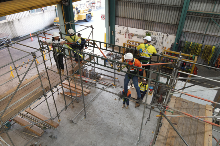 Workers equipped with safety harnesses constructing a complex scaffolding structure at height inside an industrial facility, demonstrating advanced scaffolding techniques taught in the Scaffolding Advanced course.