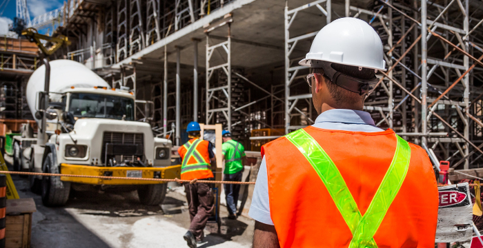 Construction worker in a safety vest and hard hat overseeing a busy construction site with scaffolding and a concrete truck in the background