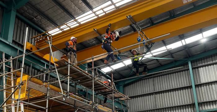 Workers wearing safety gear and harnesses performing advanced scaffolding techniques inside an industrial facility. The setup includes a complex scaffold structure and overhead rigging, demonstrating skills taught in Scaffolding Advanced training