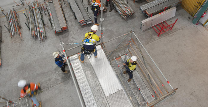 Workers in safety gear assembling a basic scaffolding structure on the ground in an indoor facility. The image captures the early stages of scaffold construction, demonstrating the foundational skills taught in Scaffolding Basic training