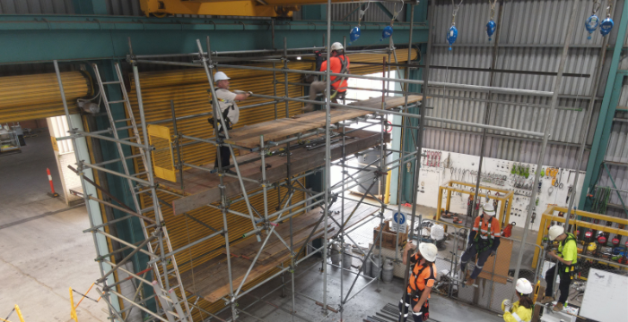 Workers wearing safety gear working on a complex scaffolding structure inside an industrial facility, demonstrating both intermediate and advanced scaffolding techniques. The image highlights the skills taught in the Scaffolding Intermediate and Advanced Combo course.