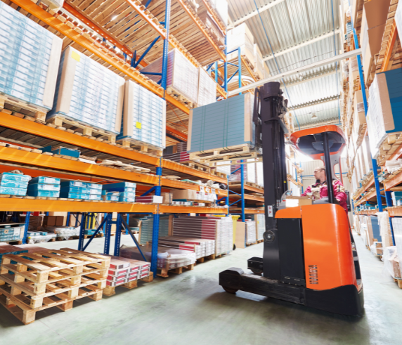 An order picker forklift in a warehouse, with the operator inside the vehicle platform. The forklift is lifting goods from high shelves, surrounded by stacked pallets and warehouse shelving, showcasing a typical order picking process